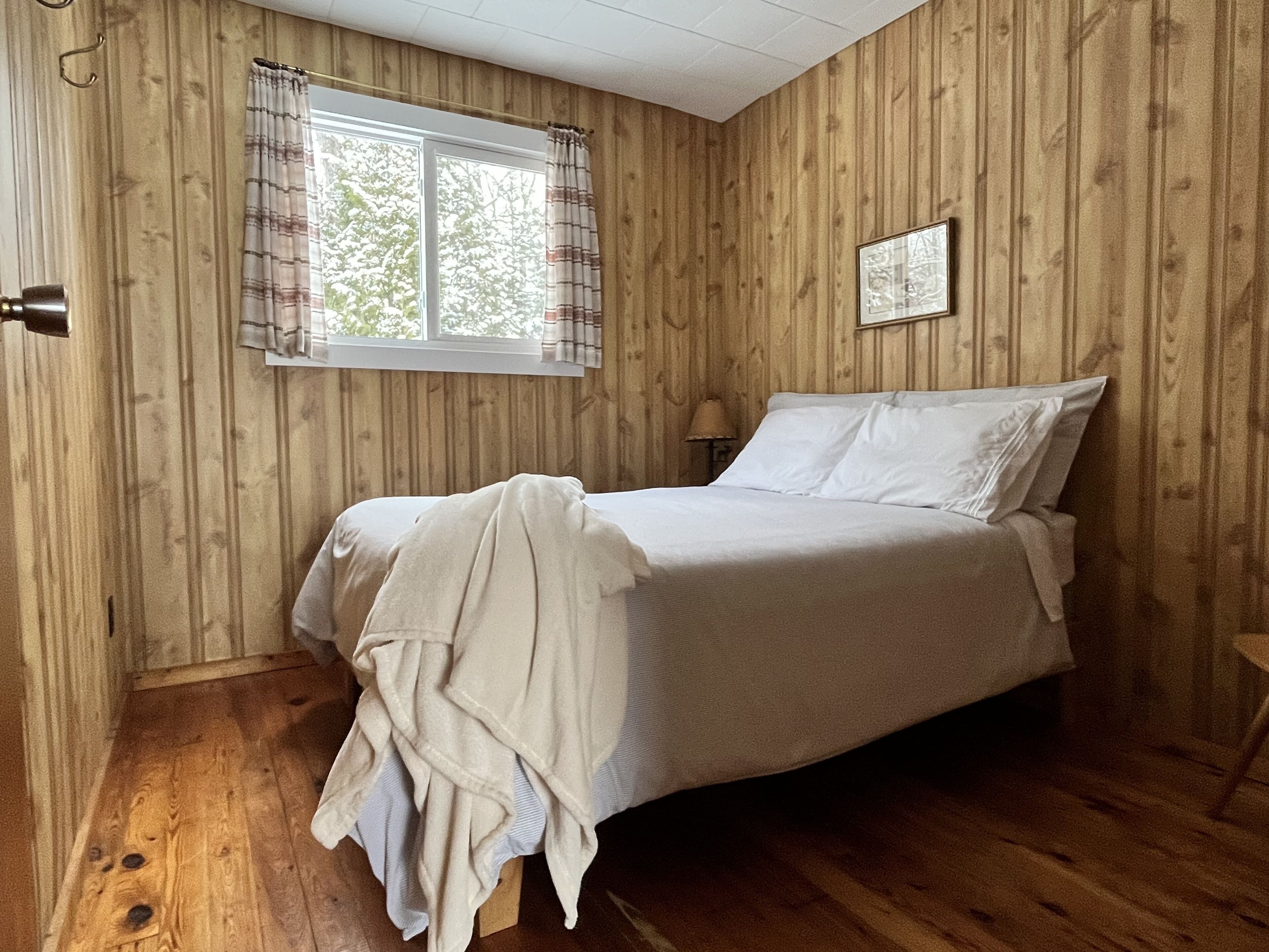 A bedroom with a wooden paneled wall, a bed with white linens, a window with curtains, and a small bedside table.