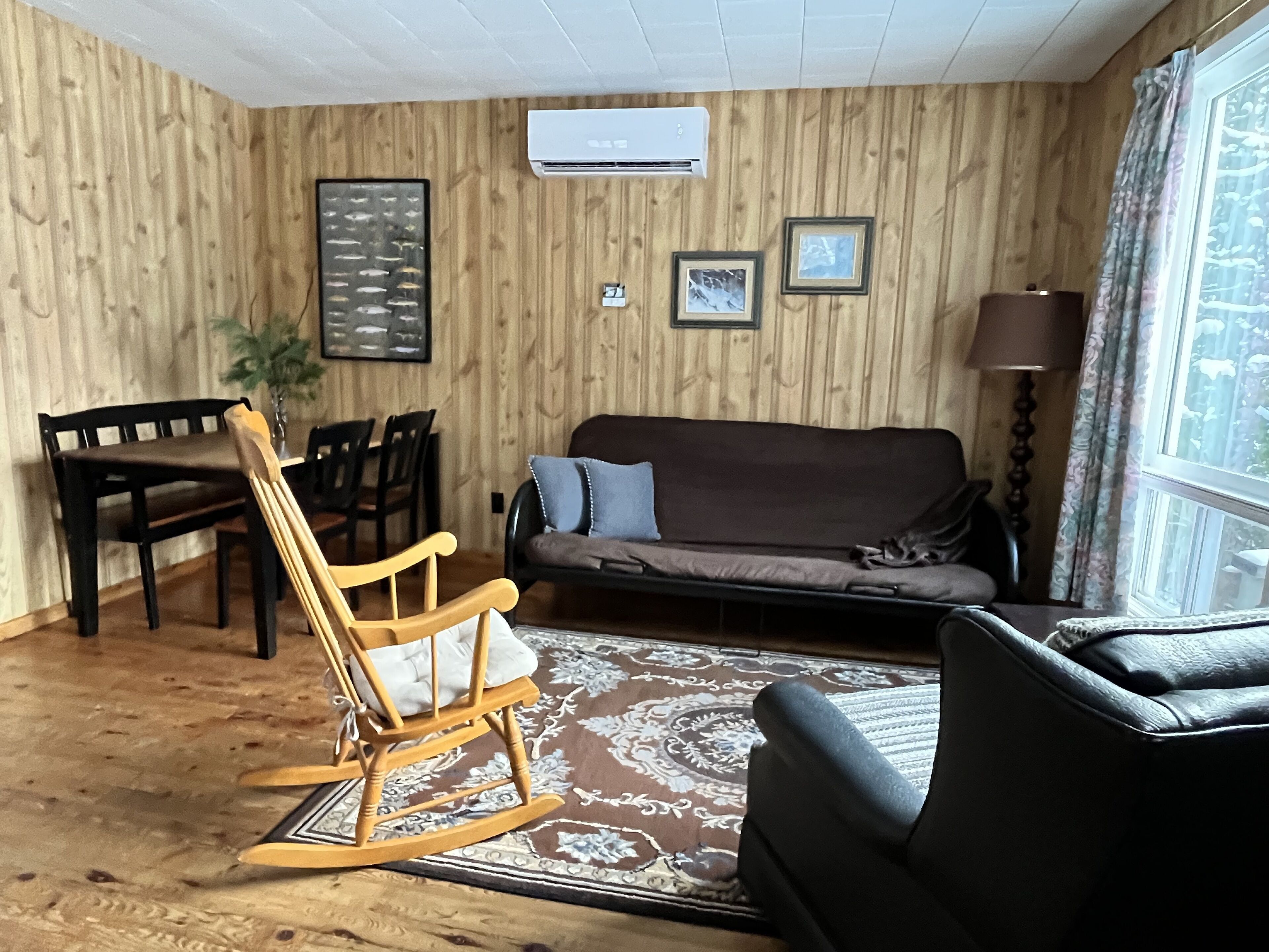 Una habitación acogedora con pared de madera, un sofá, un sillón mecedora, una mesa de comedor con sillas y una ventana con cortinas.
