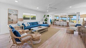 A spacious living room with a blue sofa, wicker chairs, a coffee table, and a dining area with a view of the ocean. - Oceanfront Cocoa Beach Home | Direct Beach Access | Tiki Bar & Fire Pit (Cocoa Beach)