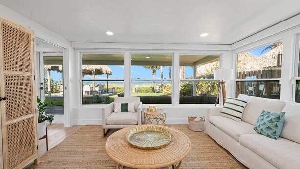 A spacious living room with a large window overlooking a tropical landscape, a white sofa, a round wicker coffee table, and a rattan door. - Oceanfront Cocoa Beach Home | Direct Beach Access | Tiki Bar & Fire Pit (Cocoa Beach)