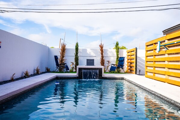 A swimming pool with a waterfall feature, surrounded by a white wall and a wooden fence.