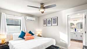 A bedroom with a bed, bedside lamps, a ceiling fan, a window with blinds, and a door leading to a bathroom.