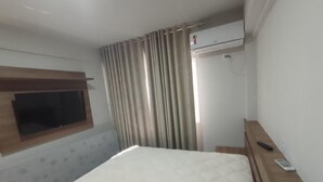 Quarto de hotel com uma cama, televisão na parede, mesa de cabeceira de madeira e janela com cortinas.