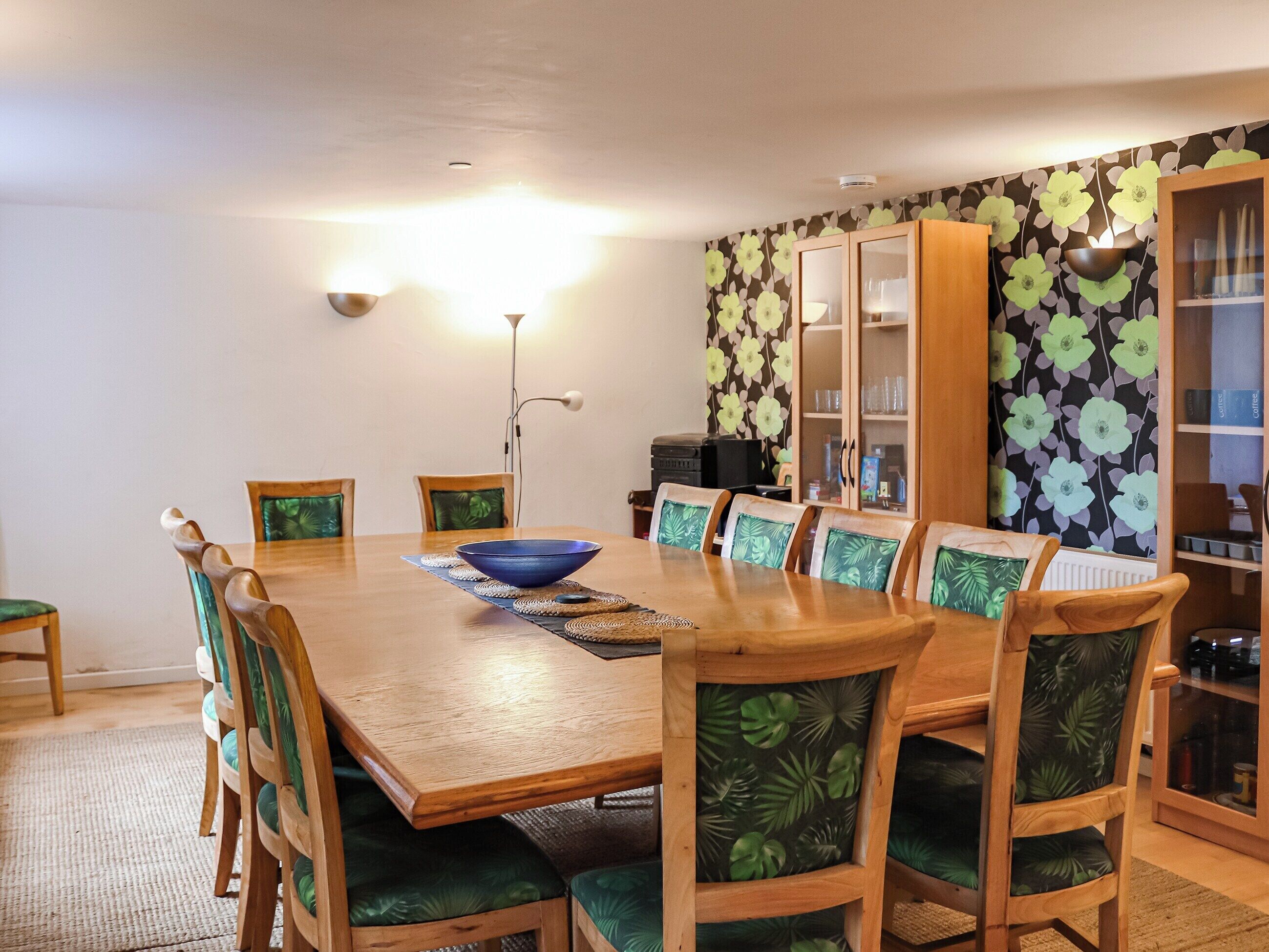 A dining area with a wooden table, green upholstered chairs, a blue bowl, and a floral wallpaper.