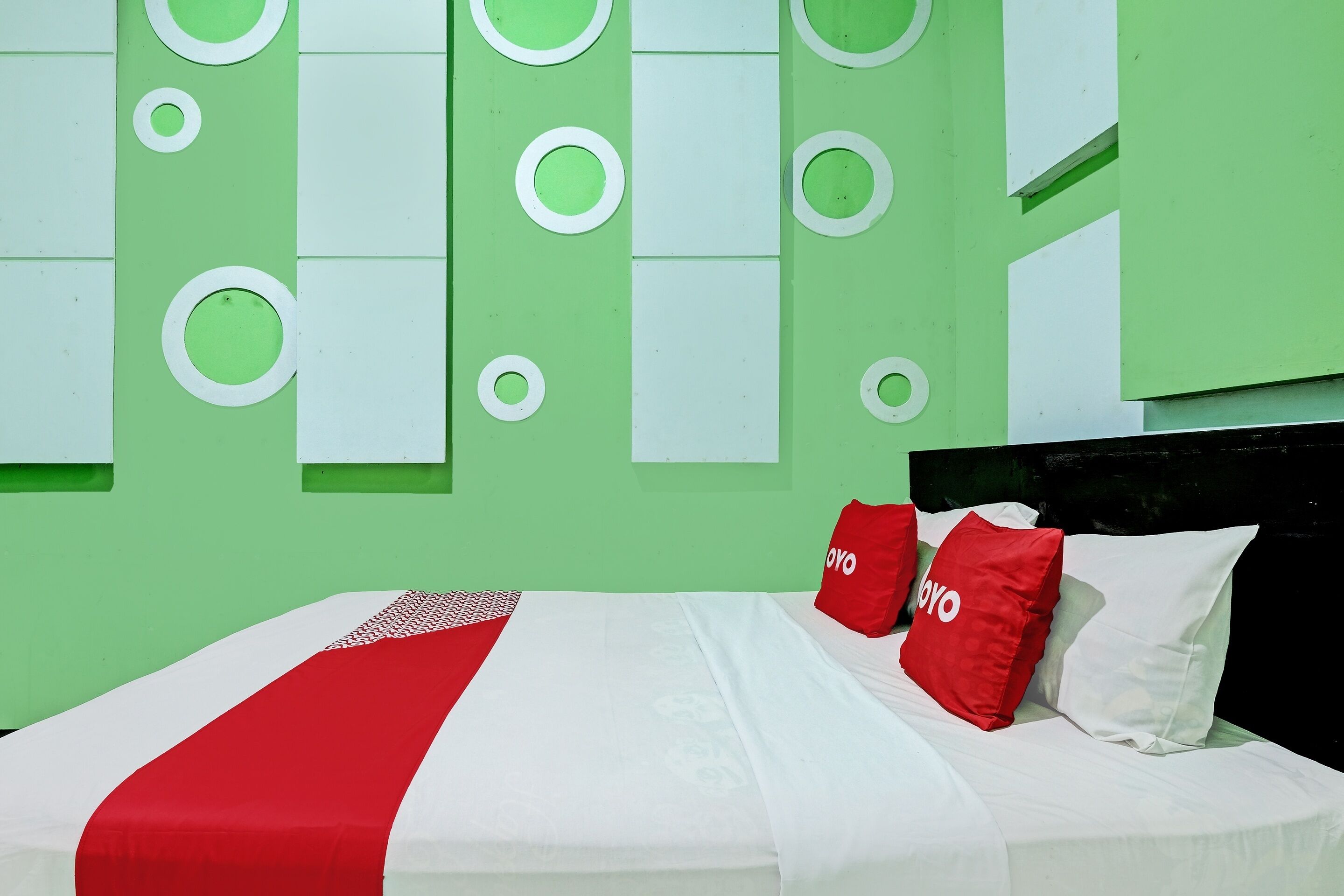 A bed with a white and red bedspread, two red pillows with "OYO" branding, and a green wall with circular and rectangular patterns.