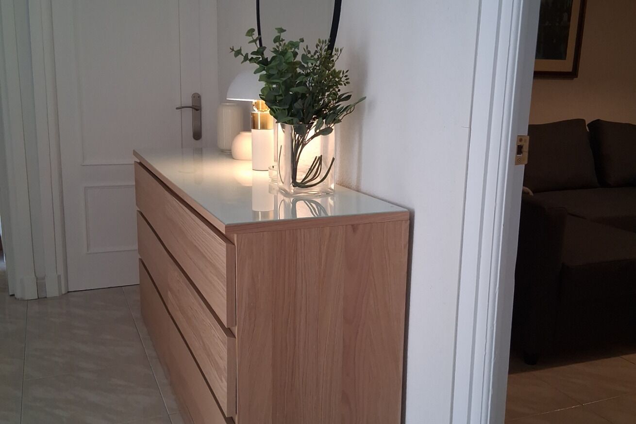 A wooden dresser with drawers, a vase with green plants, and a lamp in the background.