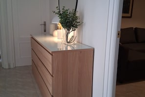 Un aparador de madera con cajones, un florero con plantas verdes y una lámpara al fondo.
