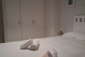 Una cama bien hecha con toallas enrolladas, una lámpara de noche y un cabecero blanco.
