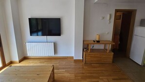 Ein Zimmer mit einem großen Flachbildfernseher an der Wand, Holzboden, einem weißen Heizkörper, einem kleinen Holztisch mit einem Korb und einer Tür zu einem weiteren Raum.