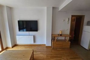 Una habitación con un televisor de pantalla plana grande montado en la pared, piso de madera, un radiador blanco, una pequeña mesa de madera con una cesta y una puerta que da a otra habitación.