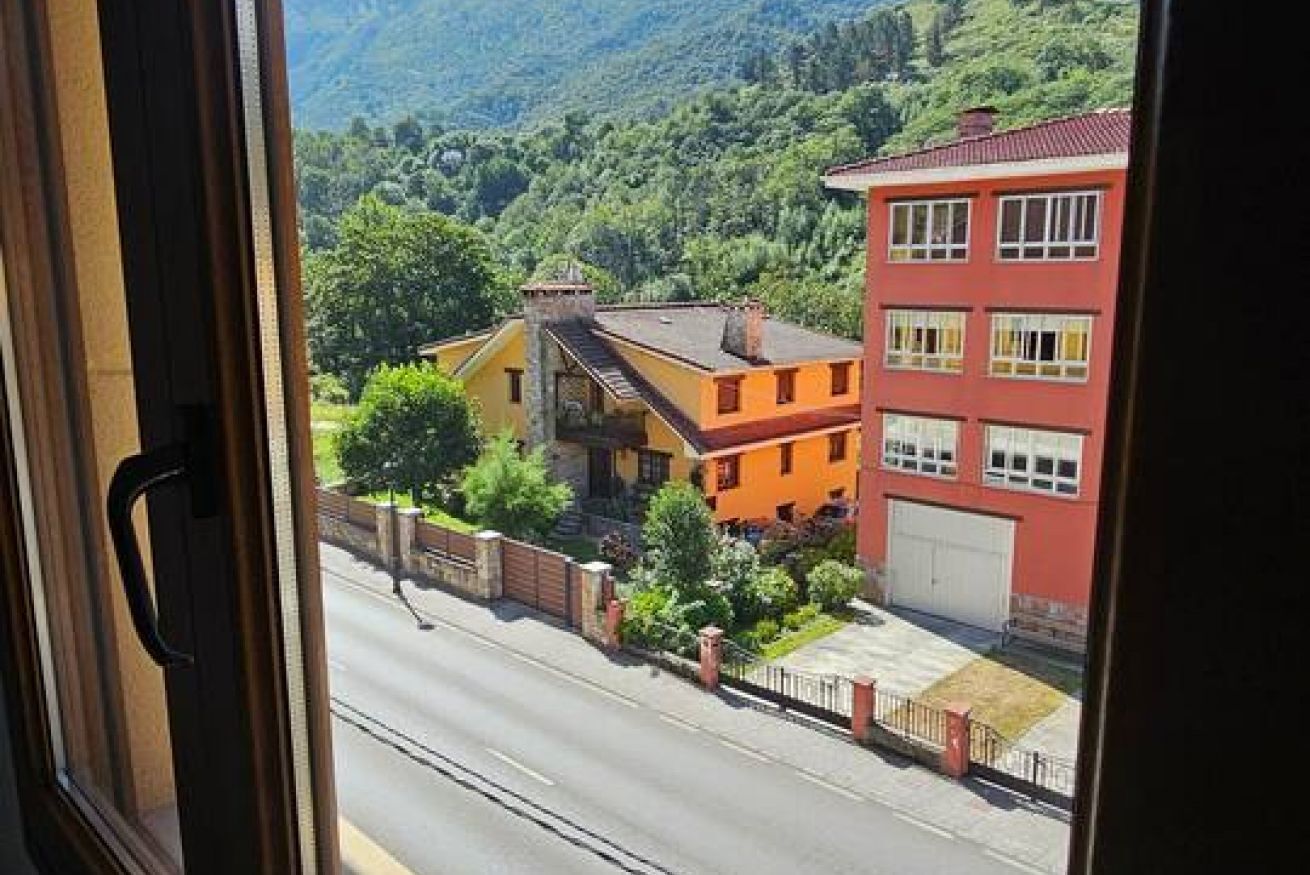 Vista a través de una ventana que muestra una calle con edificios, cielo despejado y un paisaje montañoso.