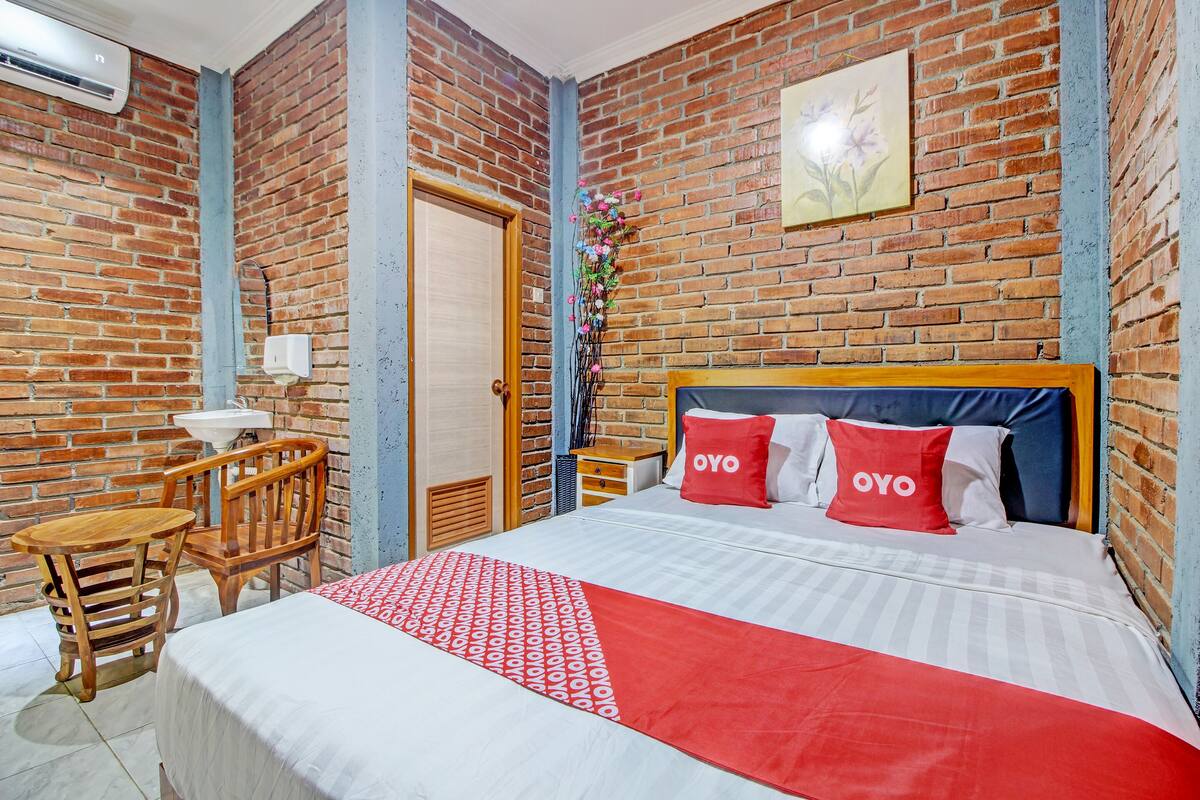 Habitación de hotel con pared de ladrillo, cama de madera con ropa de cama roja y blanca, una pequeña mesa de madera y una silla.