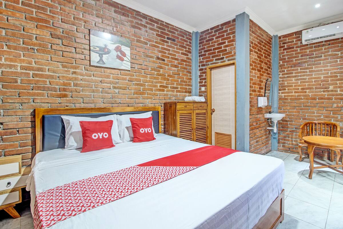 Habitación de hotel con pared de ladrillo, cama con colcha roja y blanca, cabecera de madera y silla de madera.
