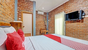Ein Hotelzimmer mit Backsteinwand, einem Bett mit weißer und roter Bettwäsche, einem Holznachttisch, einem an der Wand montierten Fernseher und einem Fenster mit Vorhängen.