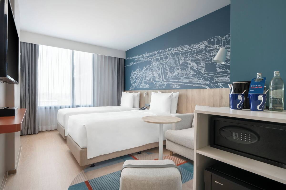 A hotel room with two beds, a desk, a small round table, and a minibar.