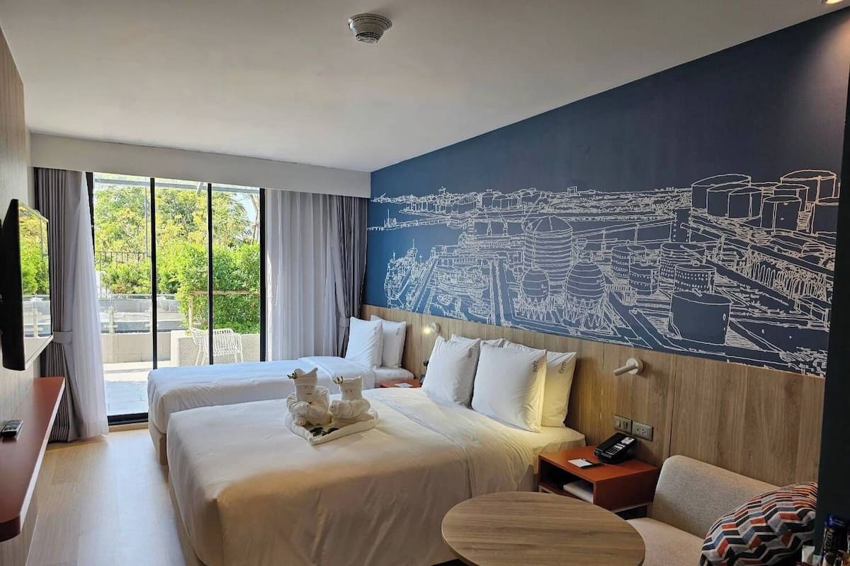 A hotel room with two beds, a large window, a cityscape mural, a small round table, and a sofa.