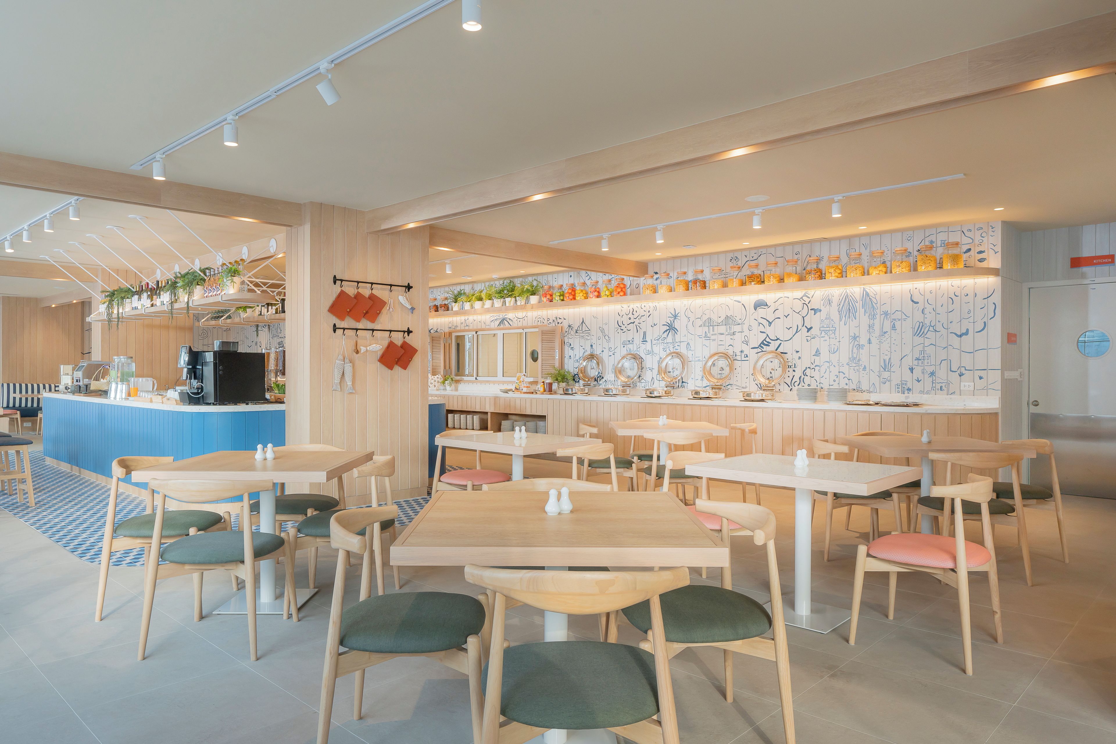 Ein modernes Café-Interieur mit Holztischen und -stühlen, einer Theke mit verschiedenen Waren sowie einer Wand mit dekorativen Fliesen.