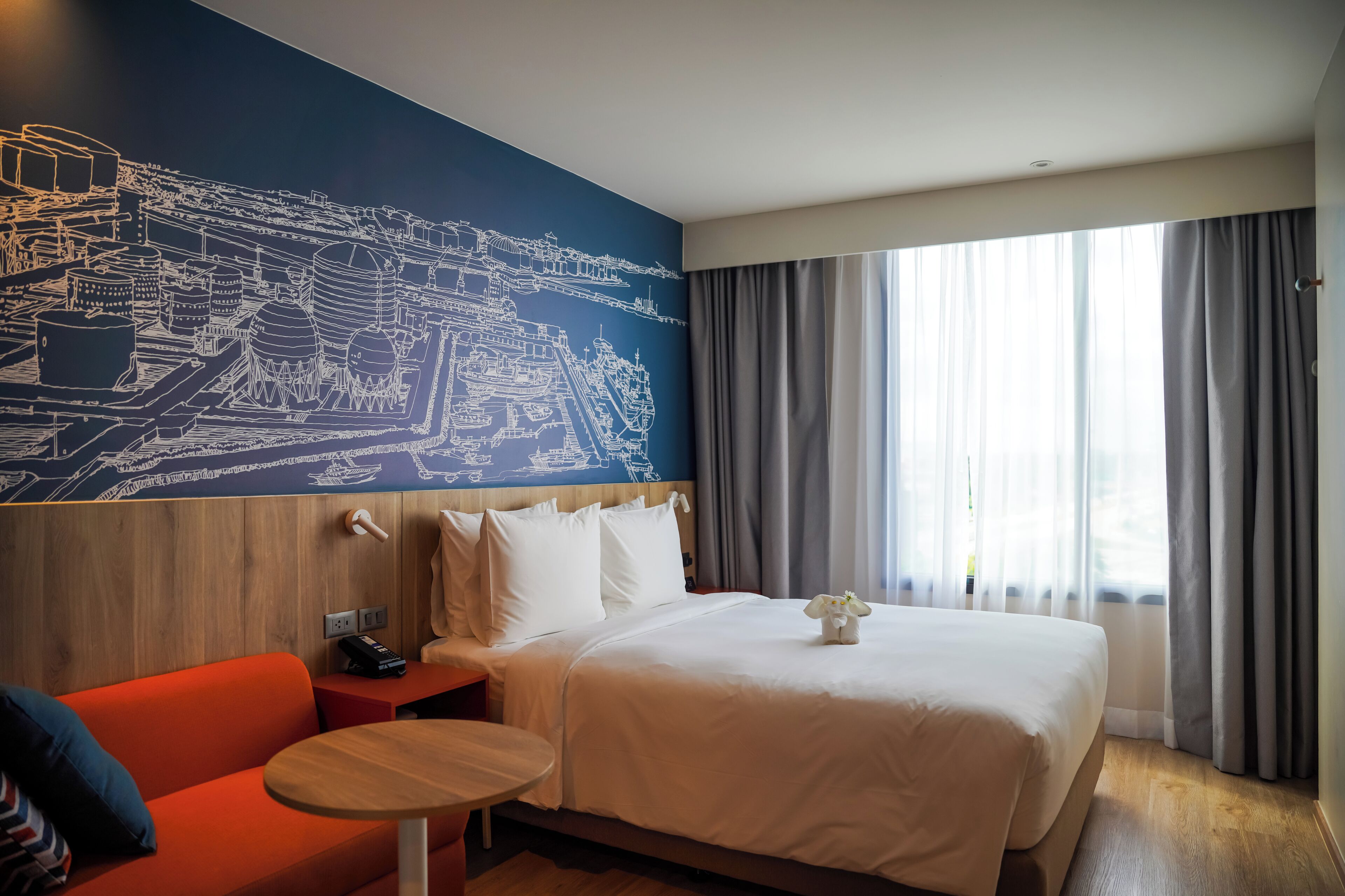Habitación de hotel con una cama grande, un sofá, una mesita, un mural en la pared y un ventanal con cortinas.