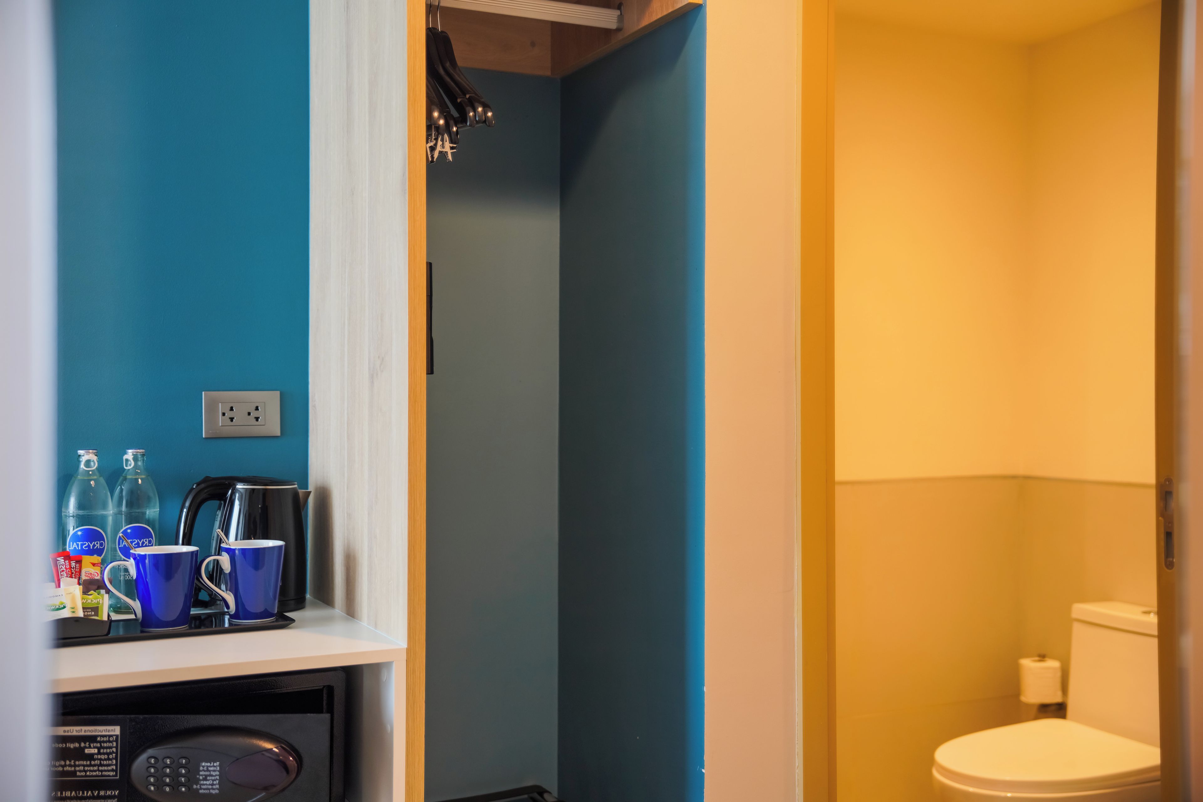 A bathroom with a toilet, a sink, and a mirror. There is a shelf with a kettle, cups, and bottles.