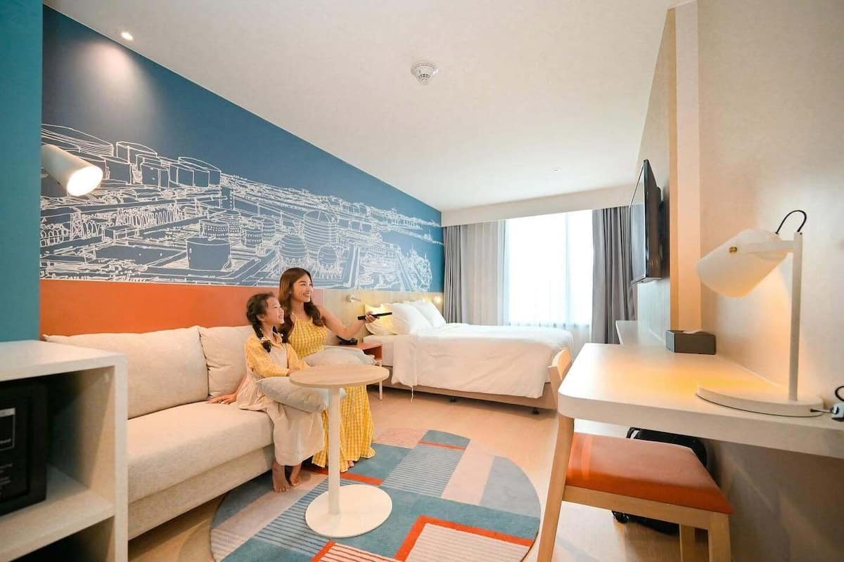 A hotel room with a bed, a sofa, a small table, a TV, and a wall mural.