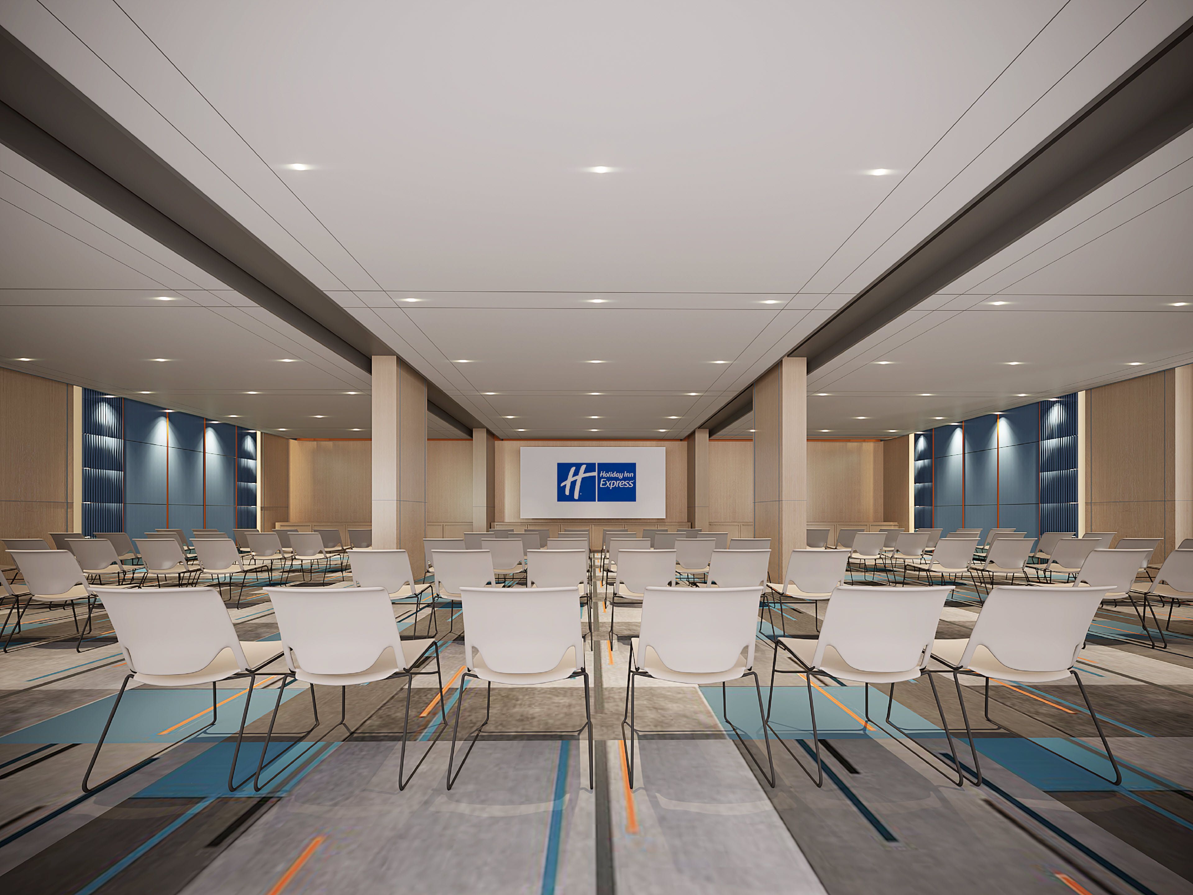 Un salón de conferencias con filas de sillas blancas, una pantalla grande mostrando el logotipo de Holiday Inn Express y un diseño de techo moderno.