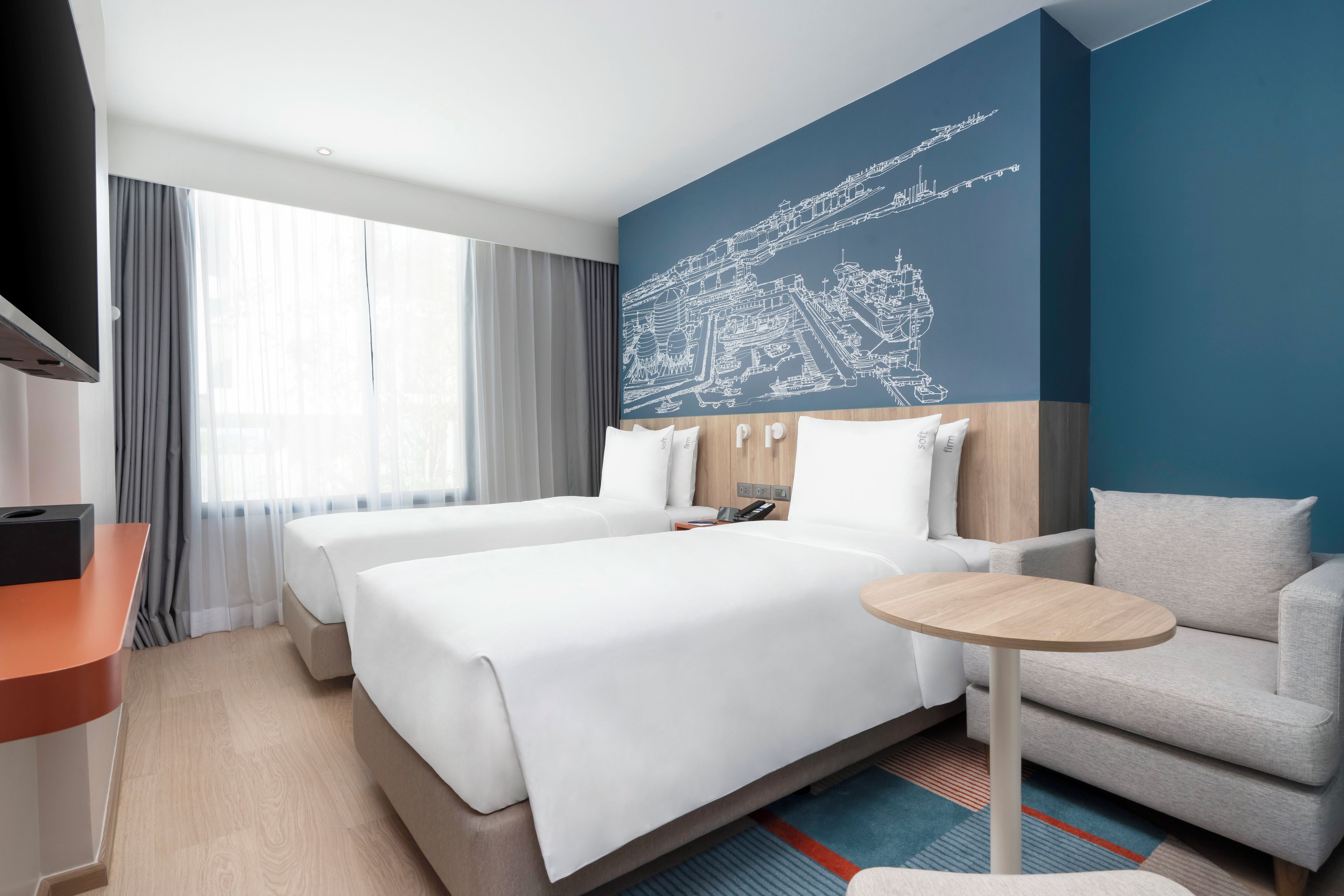 A hotel room with two beds, a TV, a desk, a chair, and a cityscape mural on the wall.