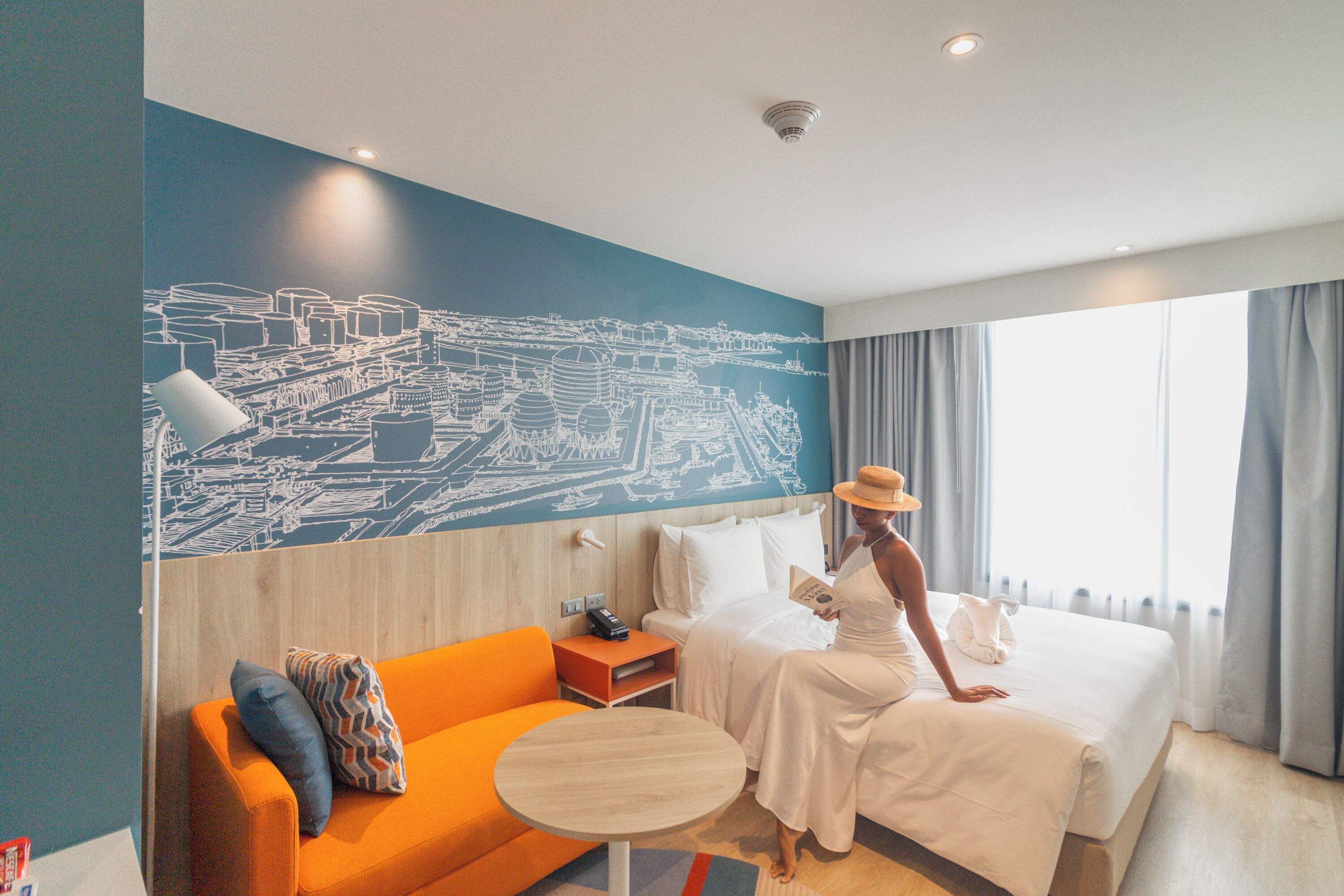 Habitación de hotel con cama, sofá, una mesita y un mural con vista urbana en la pared.