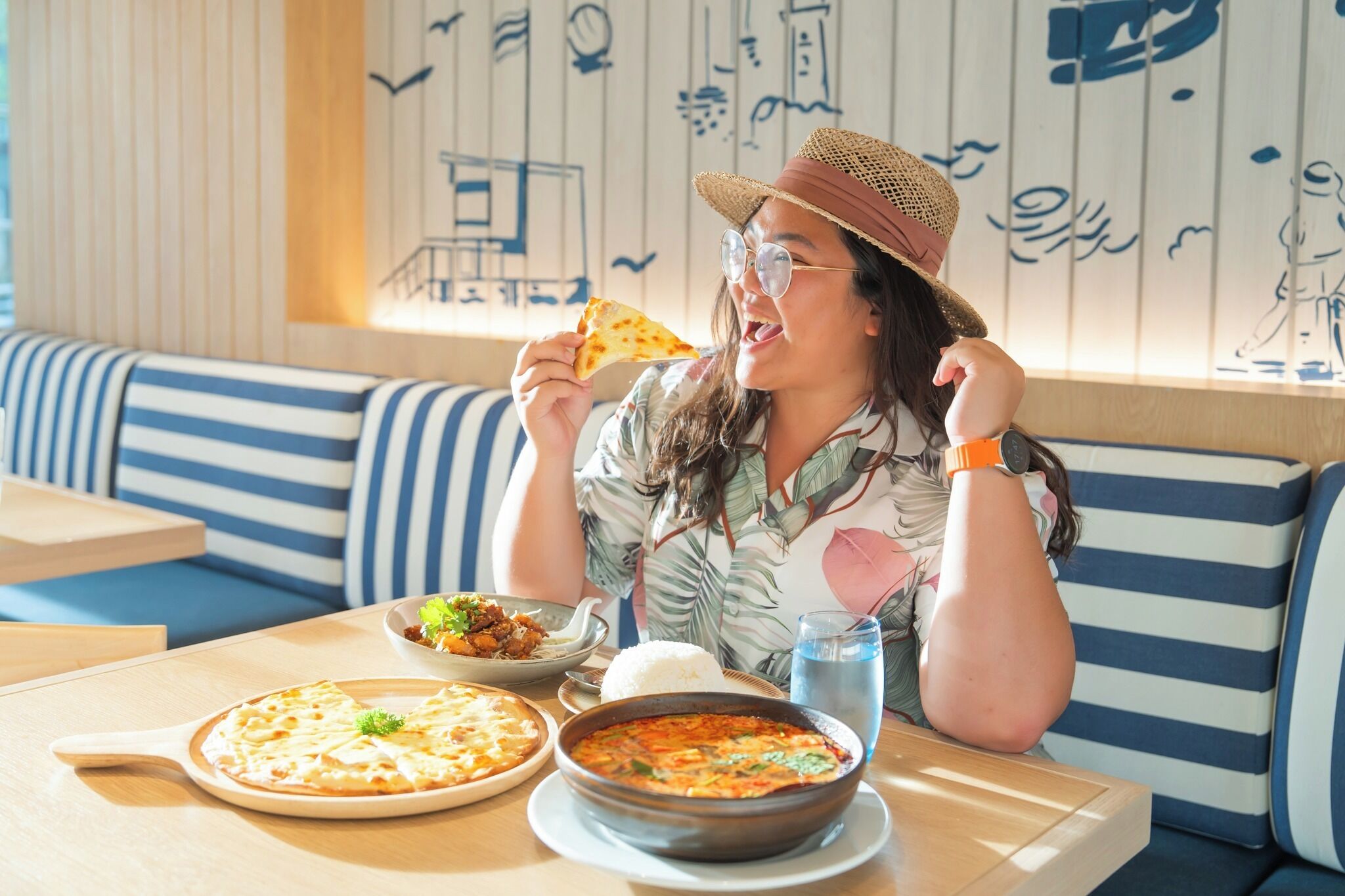 A person seated at a table with a pizza, rice, and soup. The setting includes striped blue and white cushions and a wall with blue line drawings.