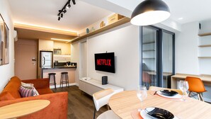 Una sala moderna con un sofá, una mesa de comedor para dos personas, una zona de cocina y un televisor que muestra 
