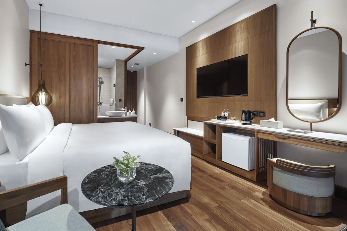 Une chambre d’hôtel moderne avec un grand lit, une table de chevet agrémentée d’un vase, un bureau en bois avec un miroir, une télévision et un petit réfrigérateur.