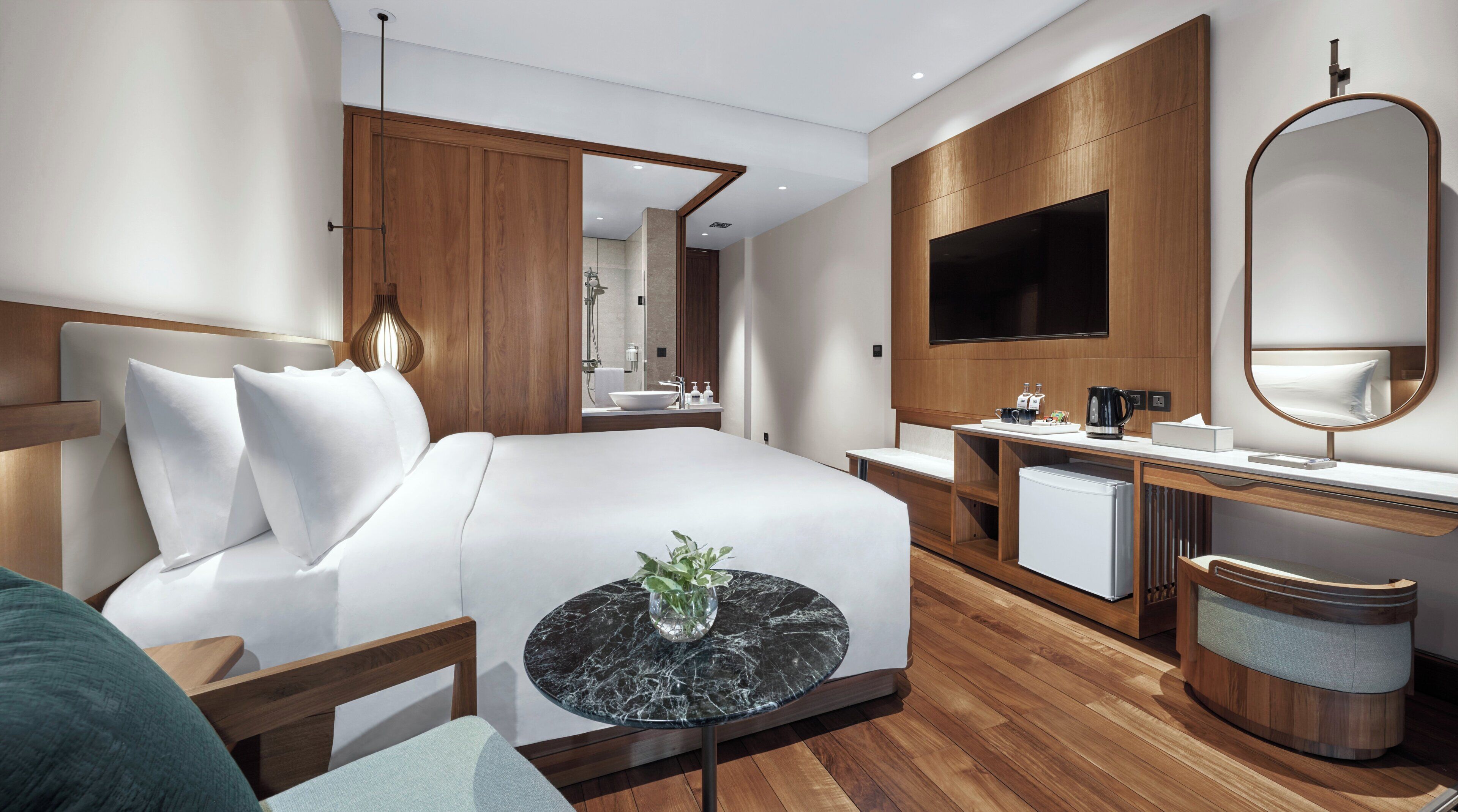 A modern hotel room with a large bed, a bedside table with a vase, a wooden desk with a mirror, a TV, and a small refrigerator.
