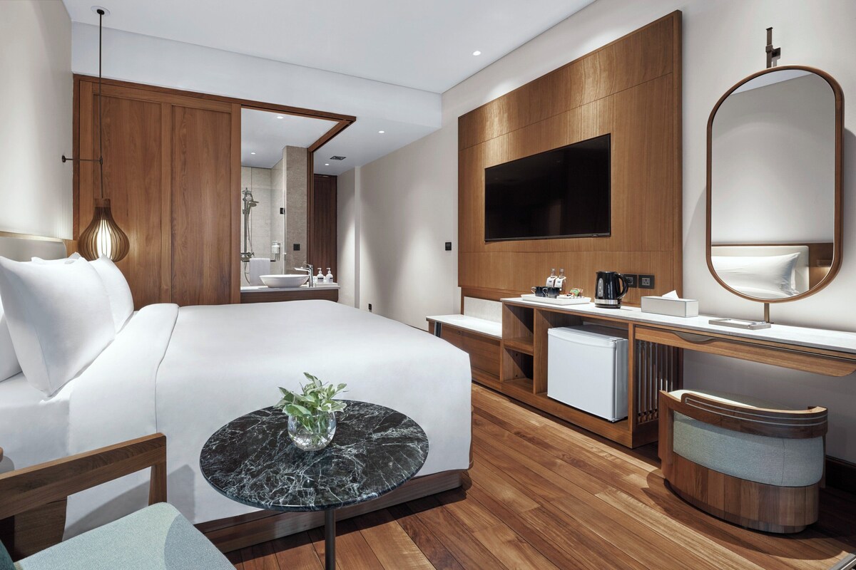 Une chambre d’hôtel moderne avec un grand lit, une table de chevet agrémentée d’un vase, un bureau en bois avec un miroir, une télévision et un petit réfrigérateur.