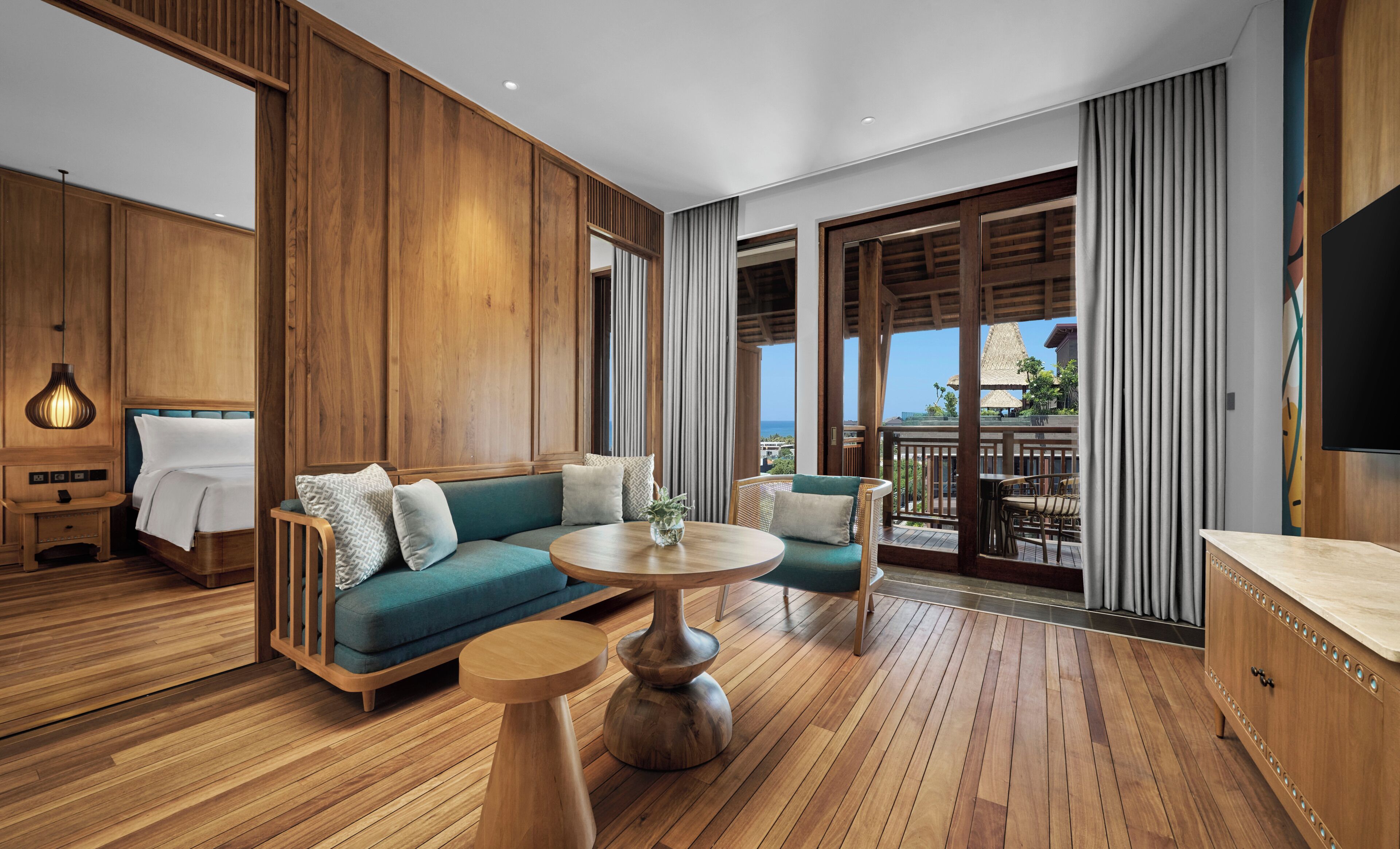 A hotel room with a wooden floor, a sofa, a round table, a TV, and a balcony with a view.
