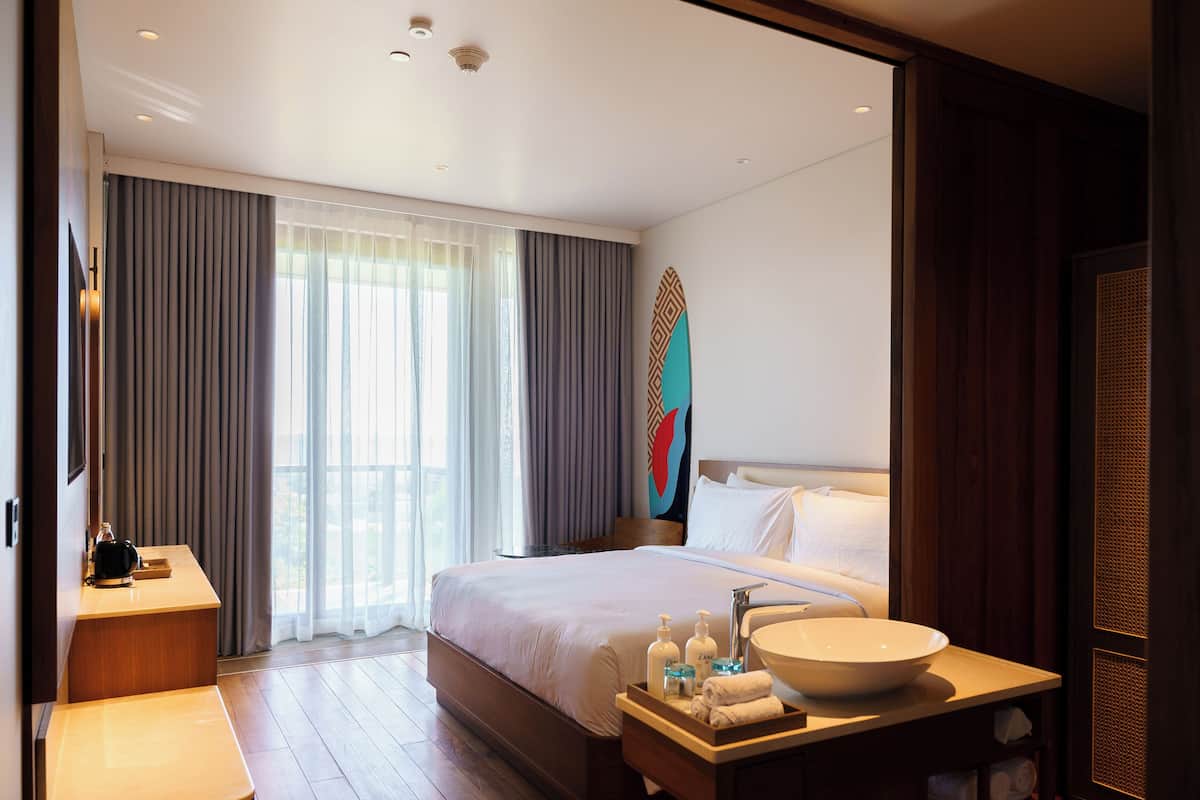 A hotel room with a large bed, a bedside table with a bowl sink, and a view of the outdoors through large windows.