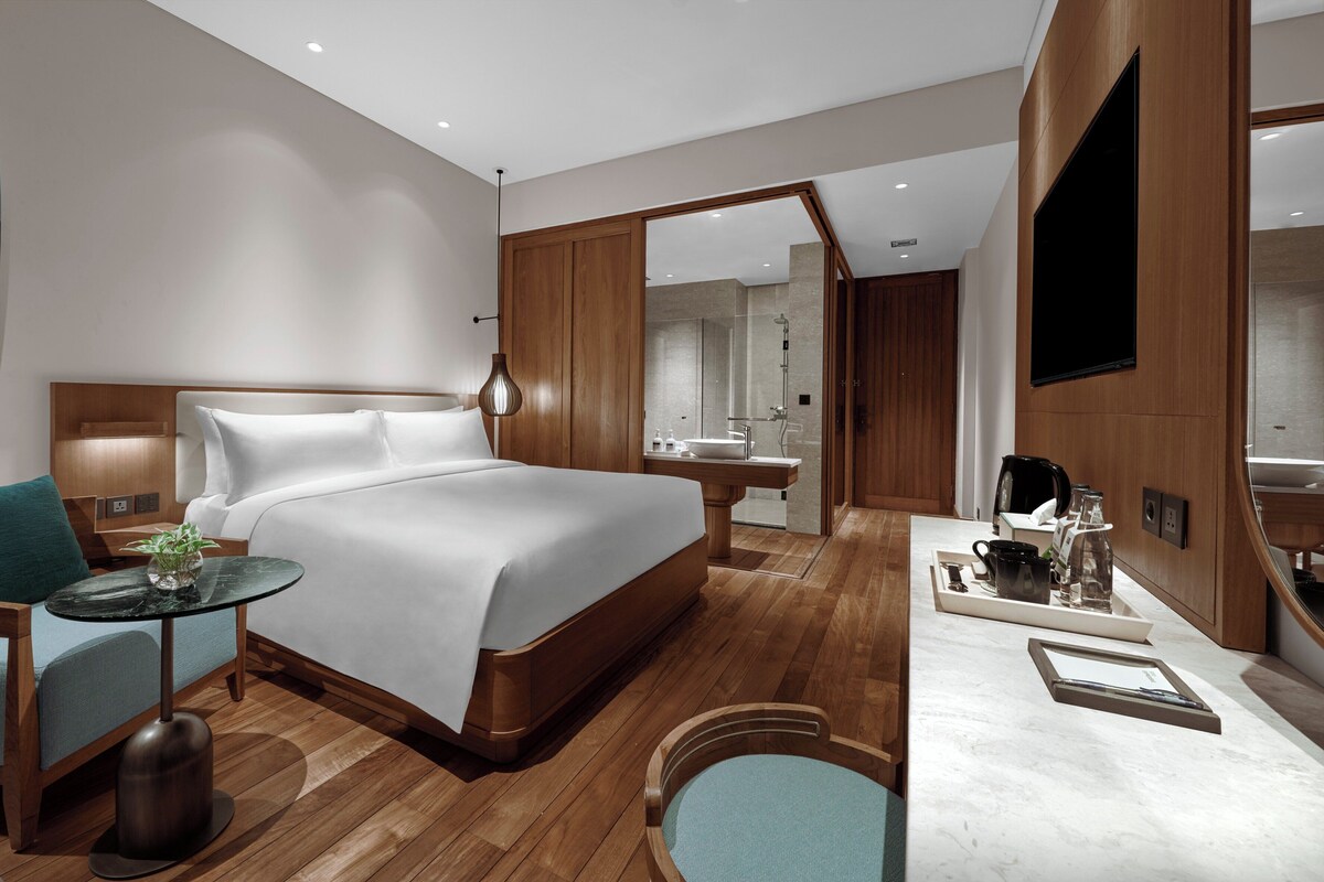 Une chambre d’hôtel moderne dotée d’un grand lit, d’un coin salon avec une table et des chaises, d’une salle de bain avec douche et lavabo, et d’un téléviseur fixé au mur.
