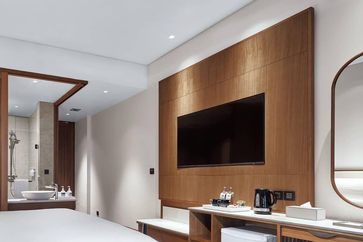 A modern hotel room with a large bed, a wooden panel wall, a flat-screen TV, a round mirror, a vanity with a sink, and a coffee maker.