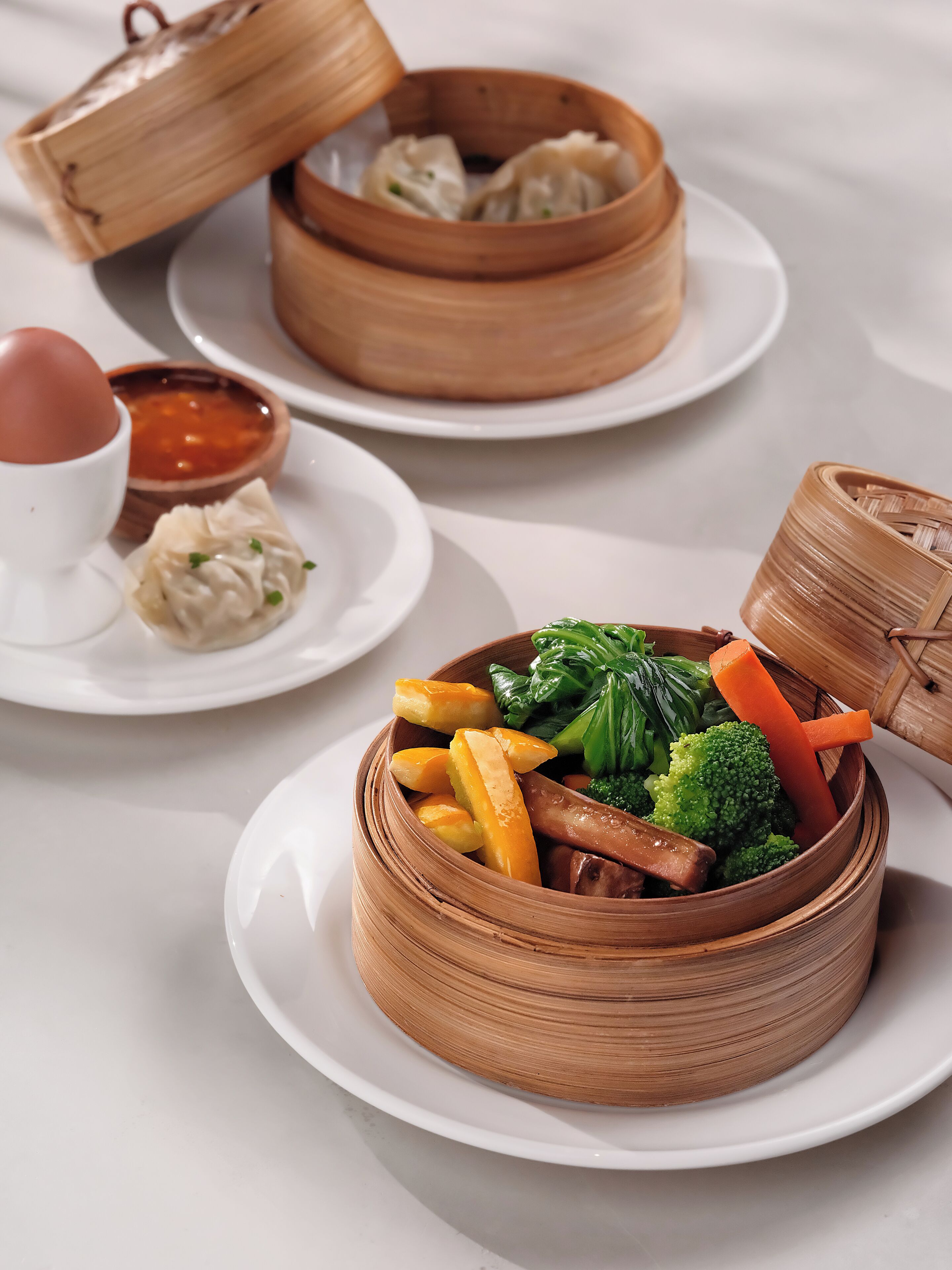 Une table avec deux paniers de cuisson à la vapeur en bambou contenant des raviolis et des légumes, ainsi qu’un petit bol de soupe.