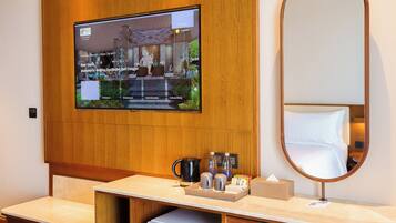 A hotel room with a wooden panel wall, a flat-screen TV mounted above a wooden desk, a round mirror, and a small refrigerator.