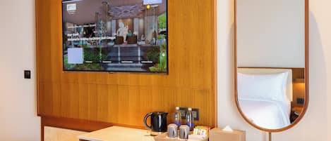 A hotel room with a wooden panel wall, a flat-screen TV mounted above a wooden desk, a round mirror, and a small refrigerator.