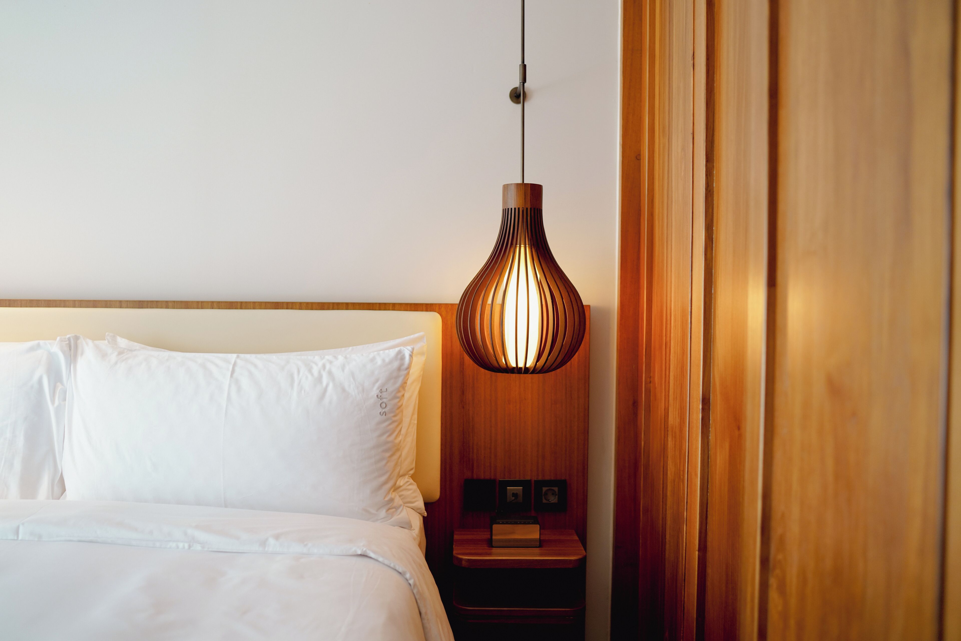 A neatly made bed with white linens and a wooden headboard, a wooden nightstand with a small clock, and a hanging pendant light.