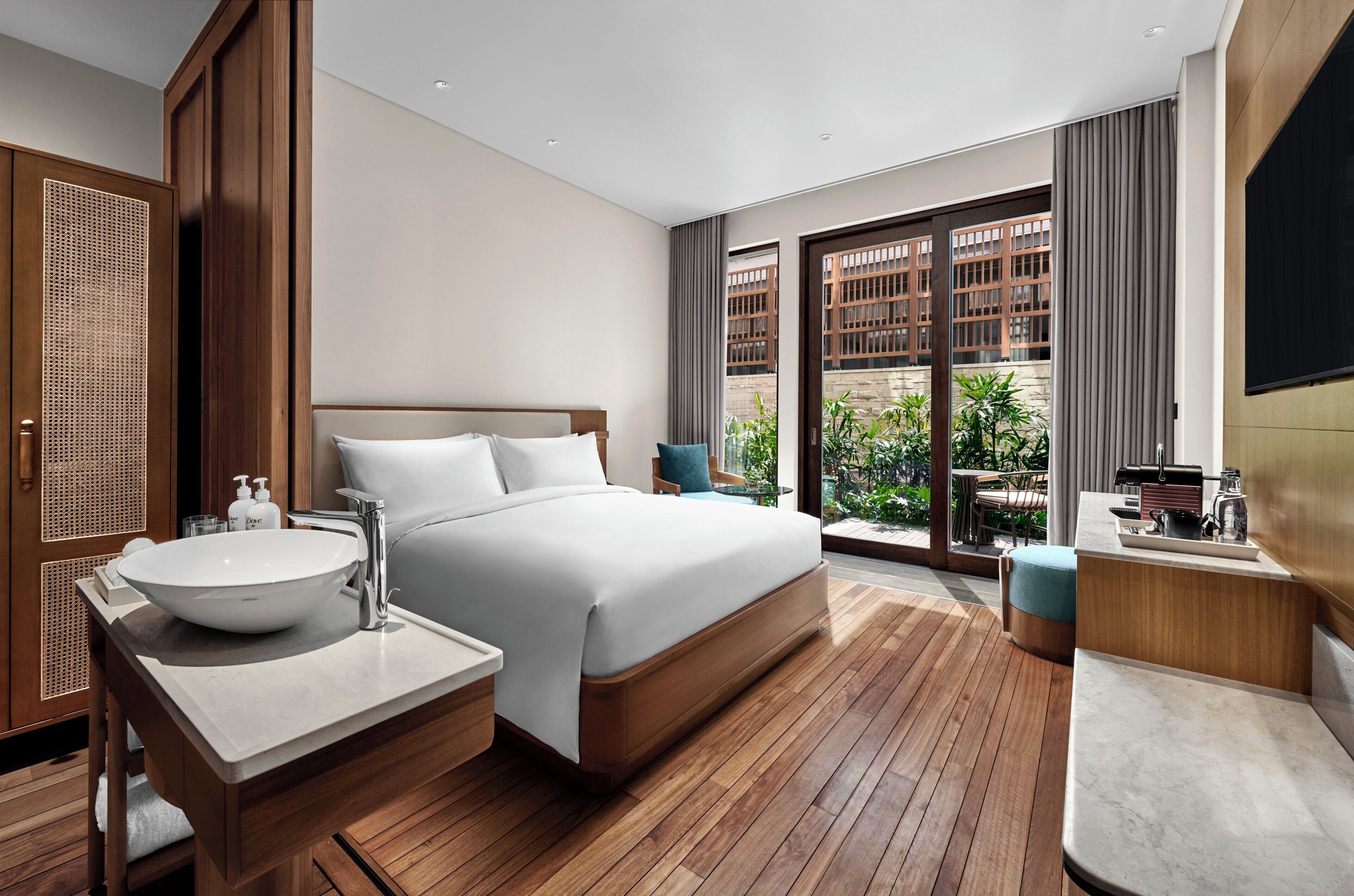 A modern hotel room with a large bed, a wooden floor, a bathroom with a sink and mirror, and a balcony with plants.