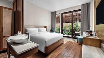 A modern hotel room with a large bed, a wooden floor, a bathroom with a sink and mirror, and a balcony with plants.
