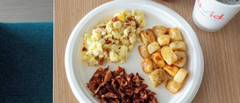A plate with scrambled eggs, roasted potatoes, and bacon, a red apple, and a cup of coffee on a wooden table.