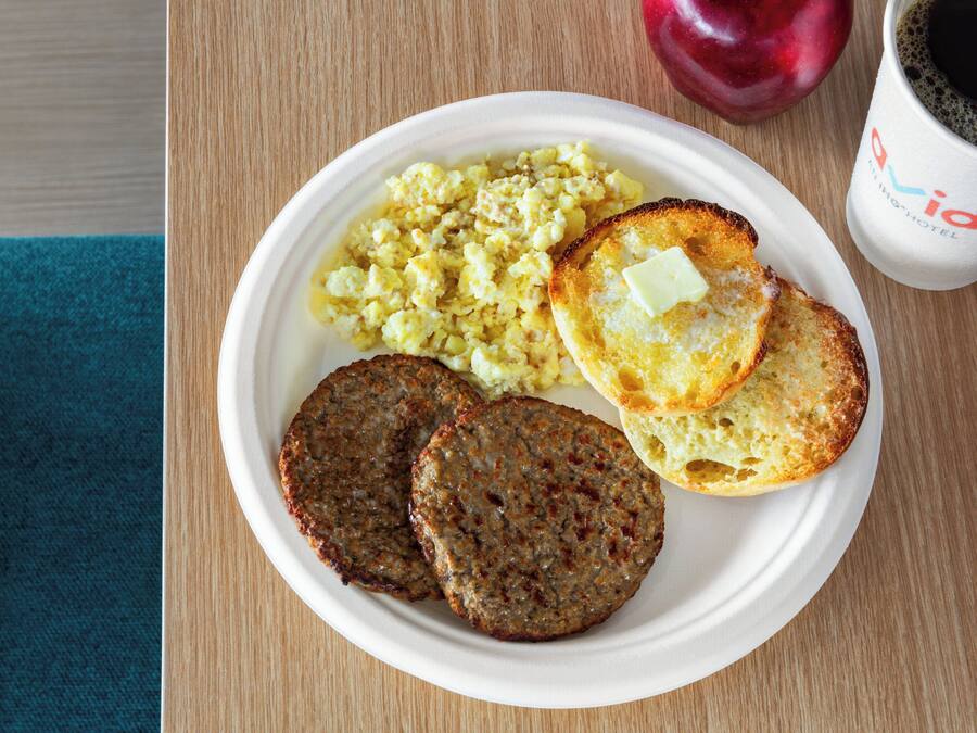 A plate with scrambled eggs, toast with butter, and two patties, alongside a coffee cup and a red apple on a wooden table.