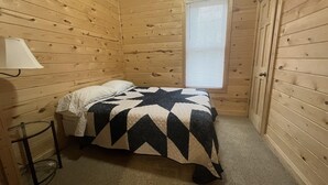 Habitació petita i acollidora amb parets de fusta, un llit individual amb un estovat estampat, una finestra amb persianes i una làmpada sobre una tauleta de nit.