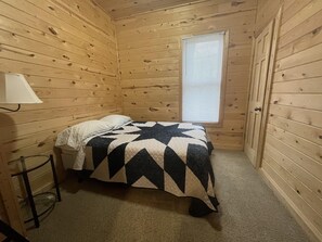 Ett litet, ombonat rum med träväggar, en enkel säng med ett mönstrat täcke, ett fönster med persienner och en lampa på ett litet bord bredvid sängen.