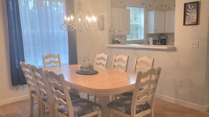 Une salle à manger avec une table ronde en bois entourée de six chaises, un lustre et une vue sur la cuisine à travers une fenêtre ornée de rideaux légers.