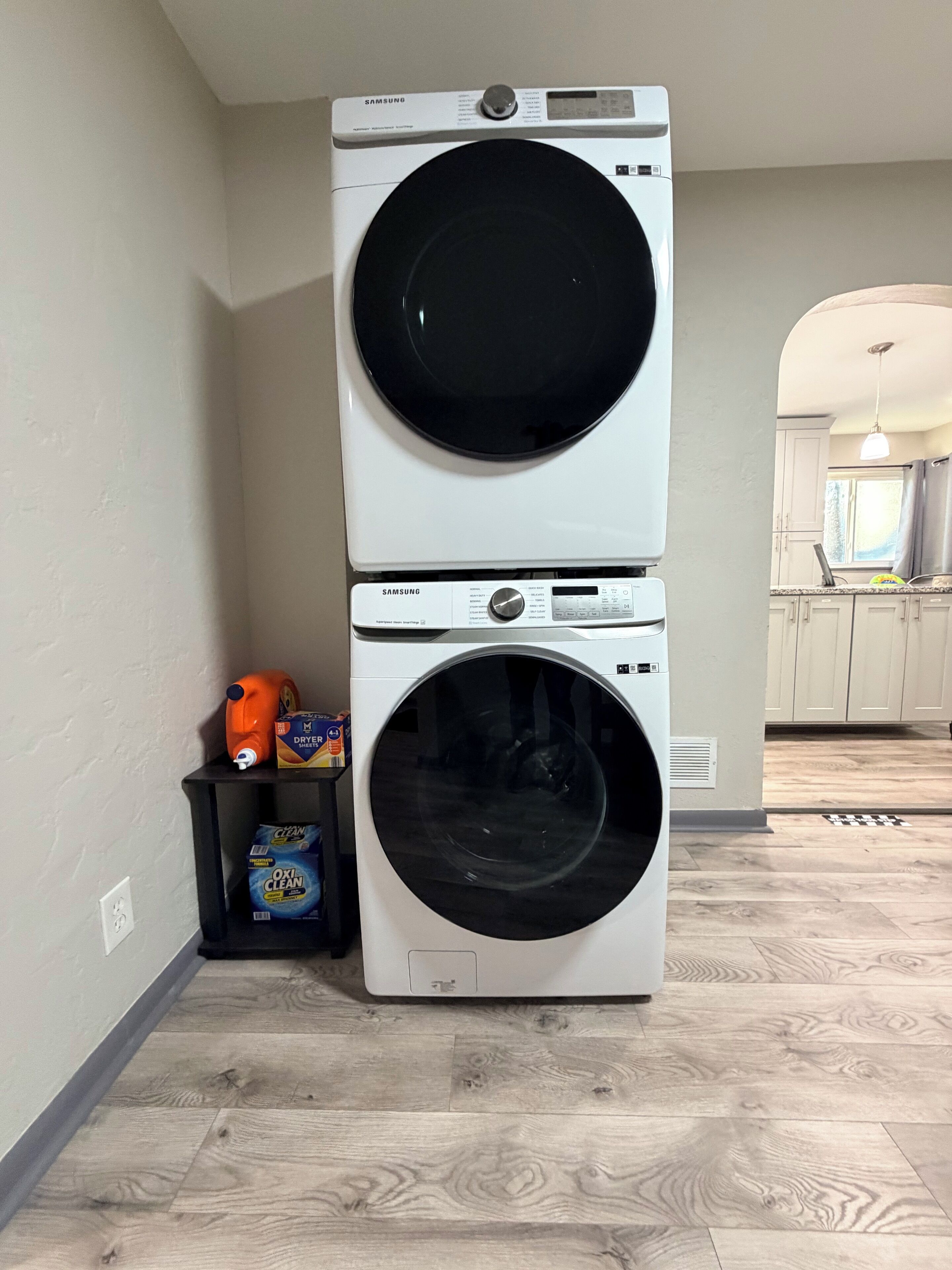 Una lavadora y secadora apiladas en un cuarto de lavado con piso de madera y una cocina visible al fondo.