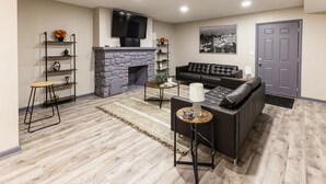 A living room with a stone fireplace, a black leather sofa, a TV mounted on the wall, and a door. - ✧KC Speedway Retreat - 10 mins to Everything!✧ (Kansas City)