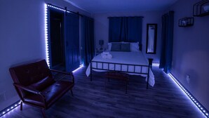 A bedroom with a bed, a chair, a bedside table, and LED strip lighting along the ceiling. - ✧KC Speedway Retreat - 10 mins to Everything!✧ (Kansas City)